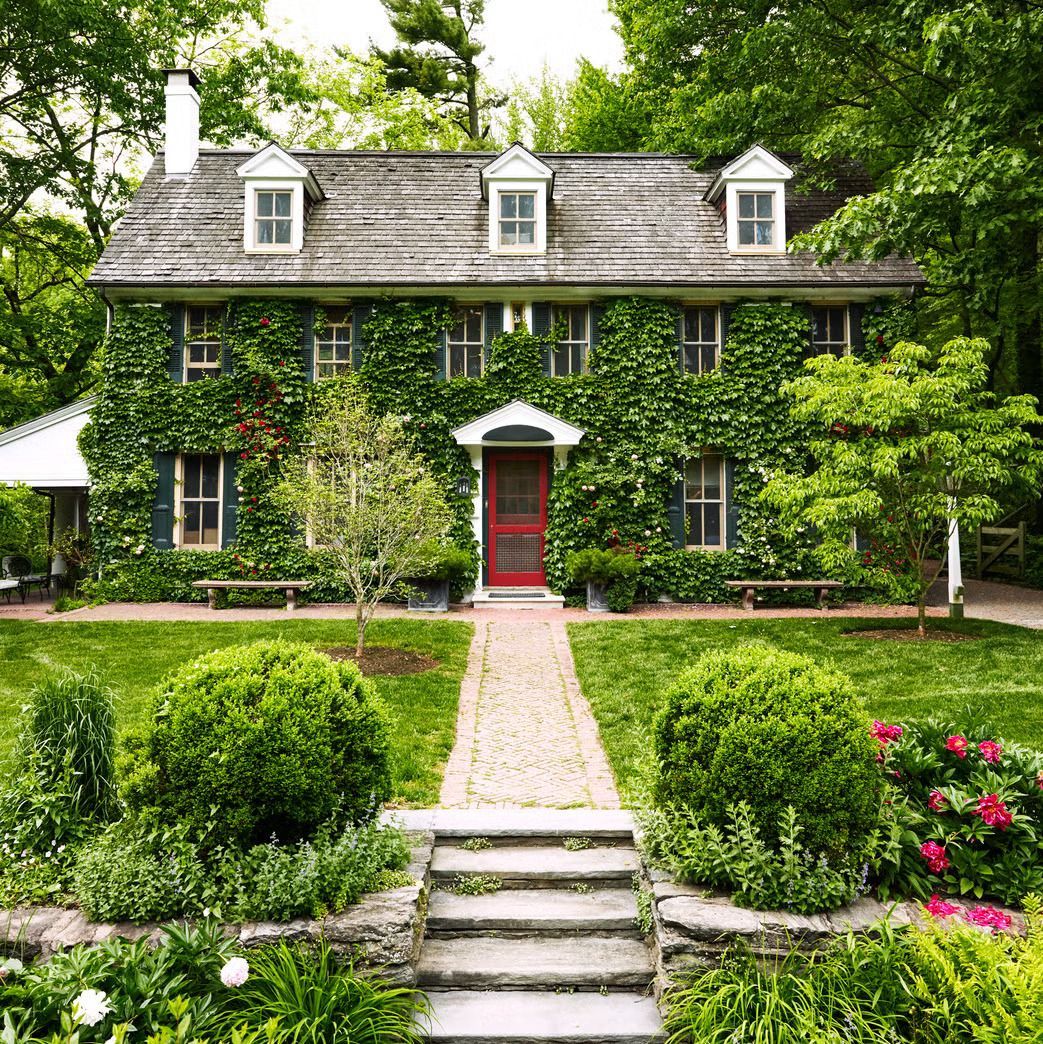 Ivy-covered two-story house with red door; stone pathway leads to entrance; lush greenery surrounds.