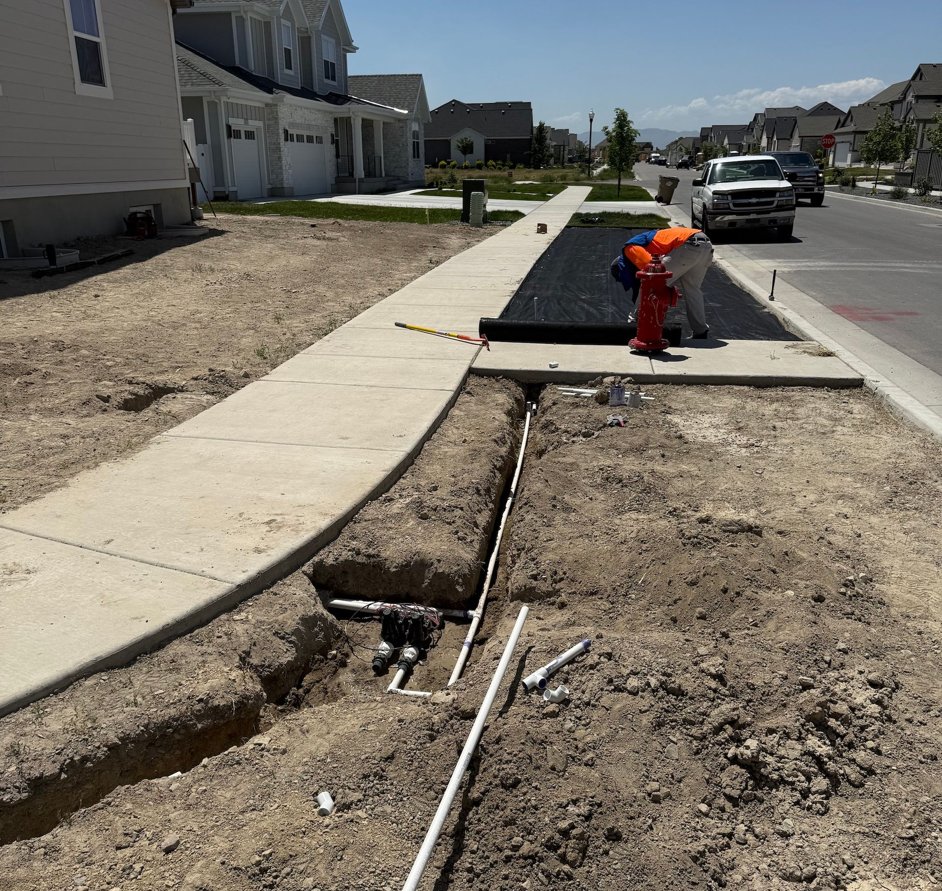 Construction site with open trenches, pipes, and sidewalk alongside a residential street. A worker is visible.