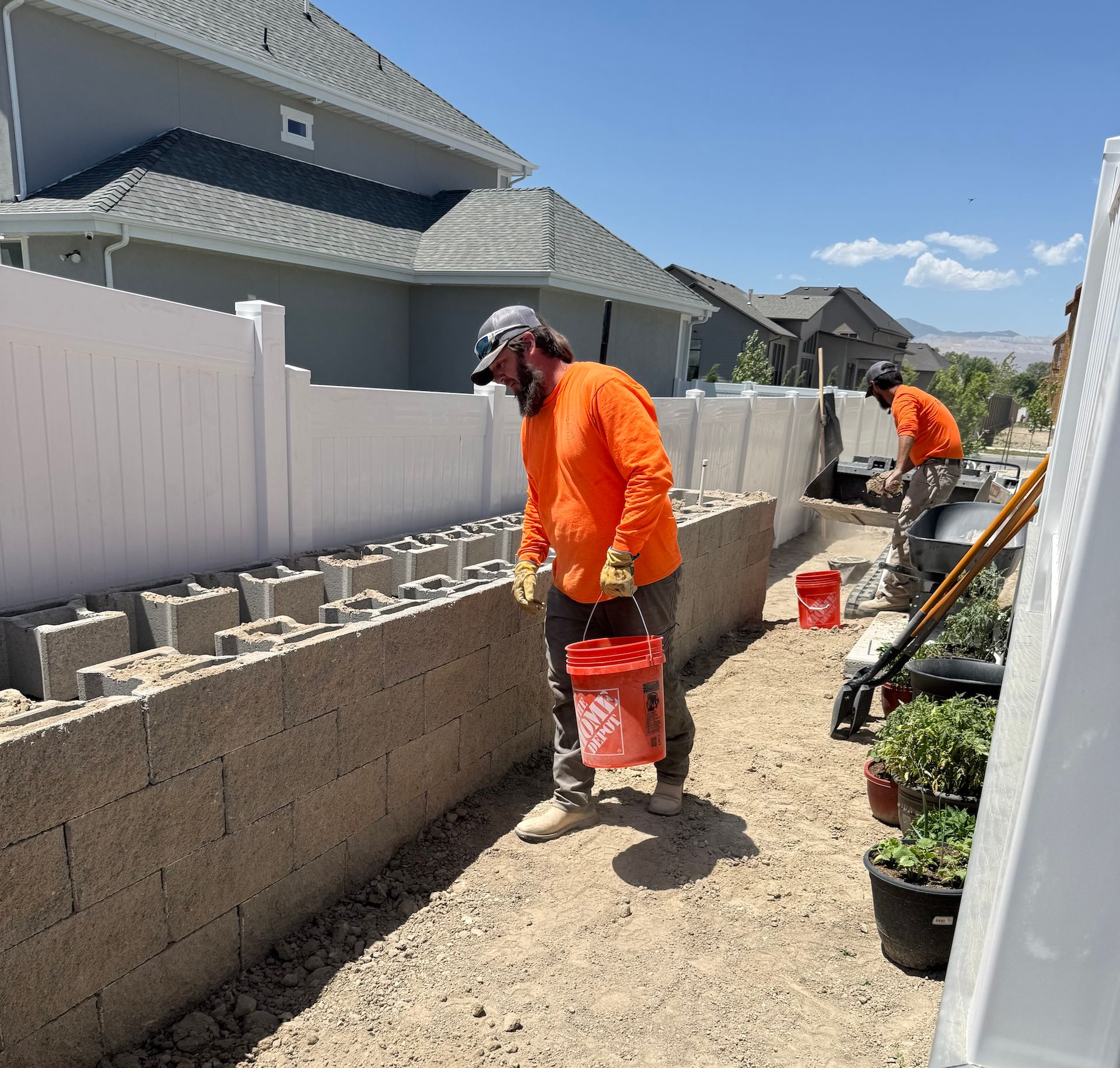 Two construction workers building a wall outdoors. One holds a red bucket. The wall is made of concrete blocks.