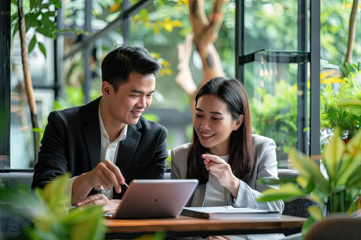 Two colleagues smiling and discussing a tablet at a café table with plants and large windows.