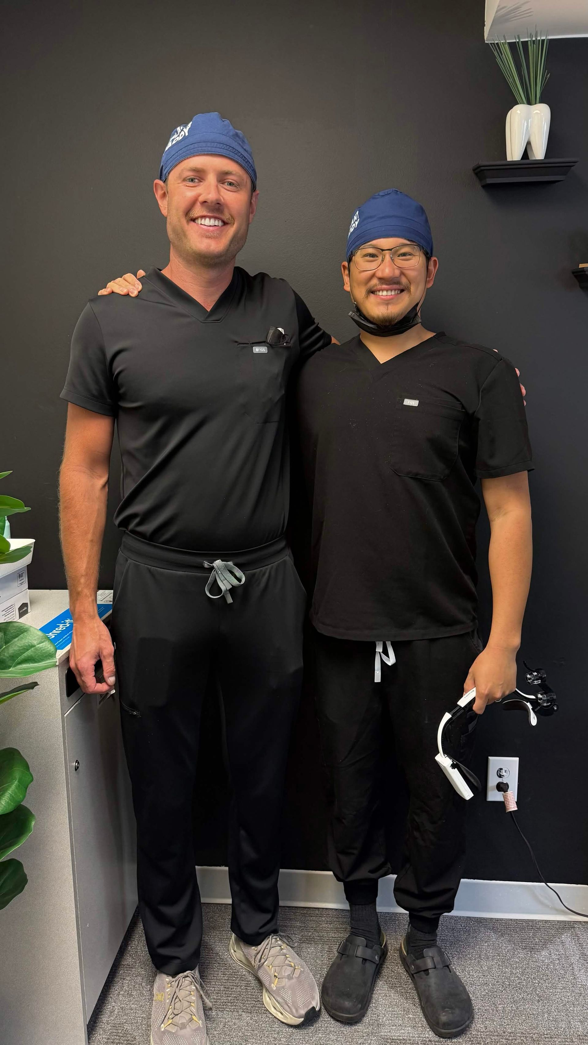 Dr. Aaron Miller and another dentist in black scrubs and blue caps stand smiling against a dark wall.