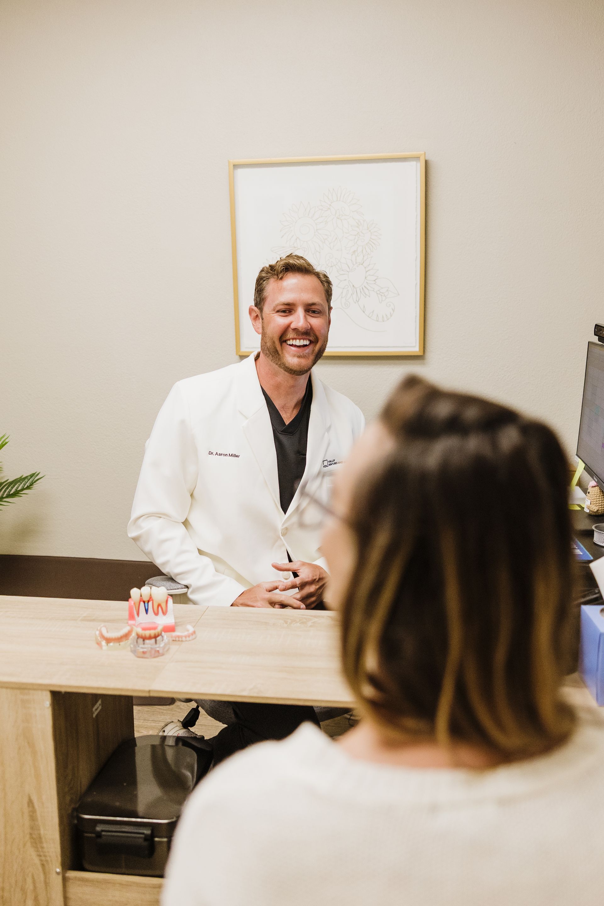 Dr. Aaron Miller in white coat talking across a desk in a clinic office, with a seated visitor in the foreground.