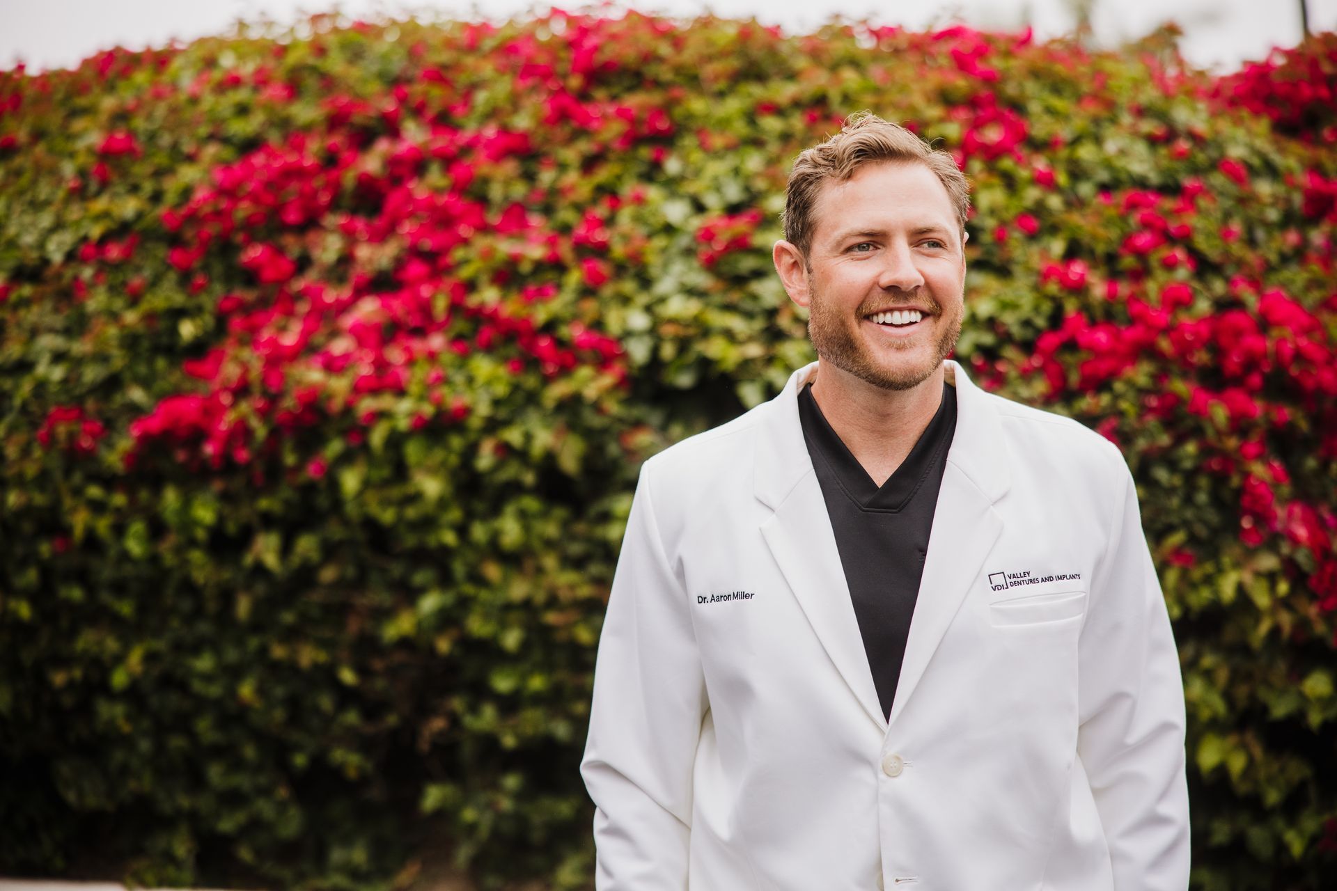 Aaron Miller smiles in a white coat standing outdoors before a pink flowering hedge