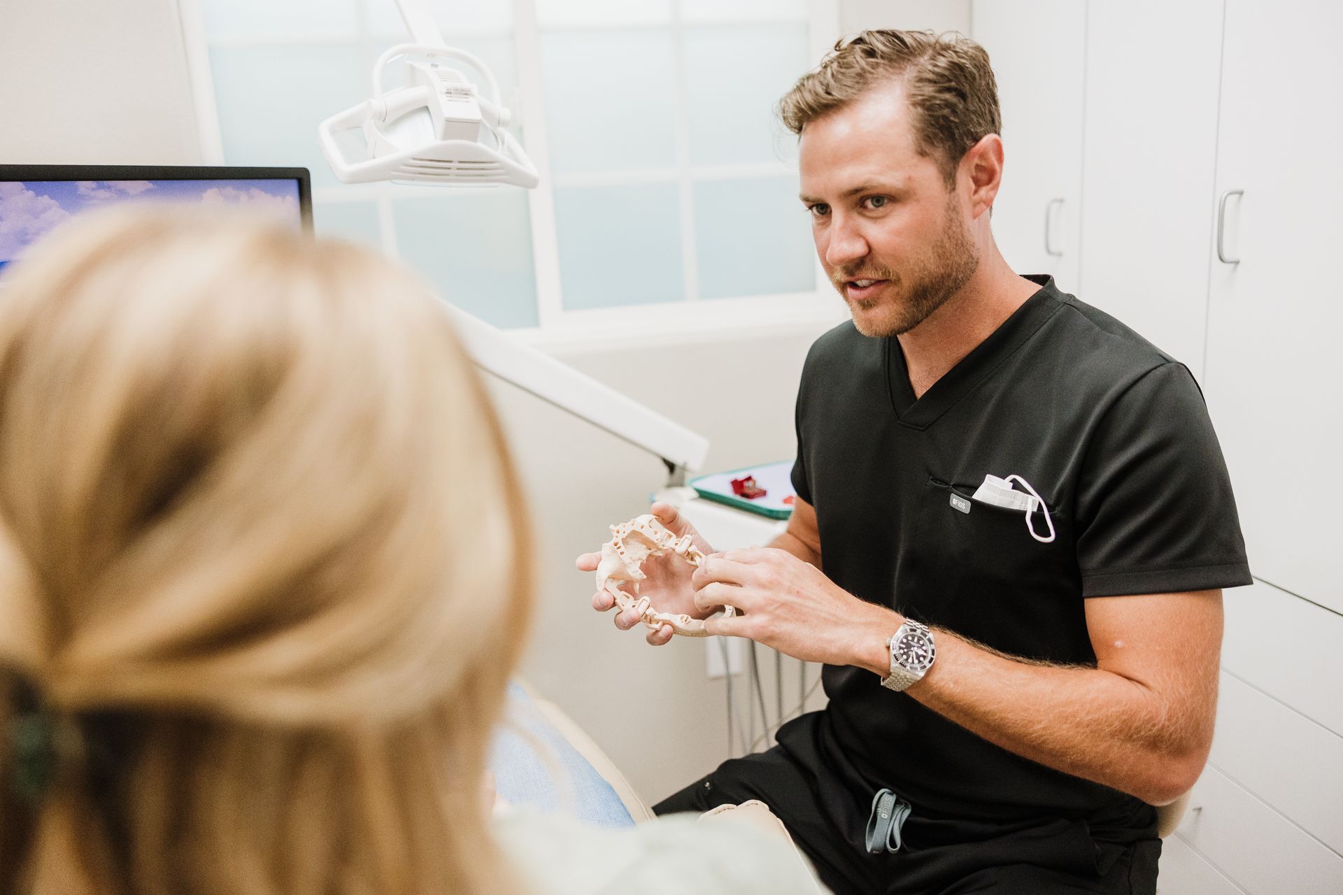 Aaron Miller sitting next to a patient in a dental office, holding a model of a jaw and explaining the model to the patient.