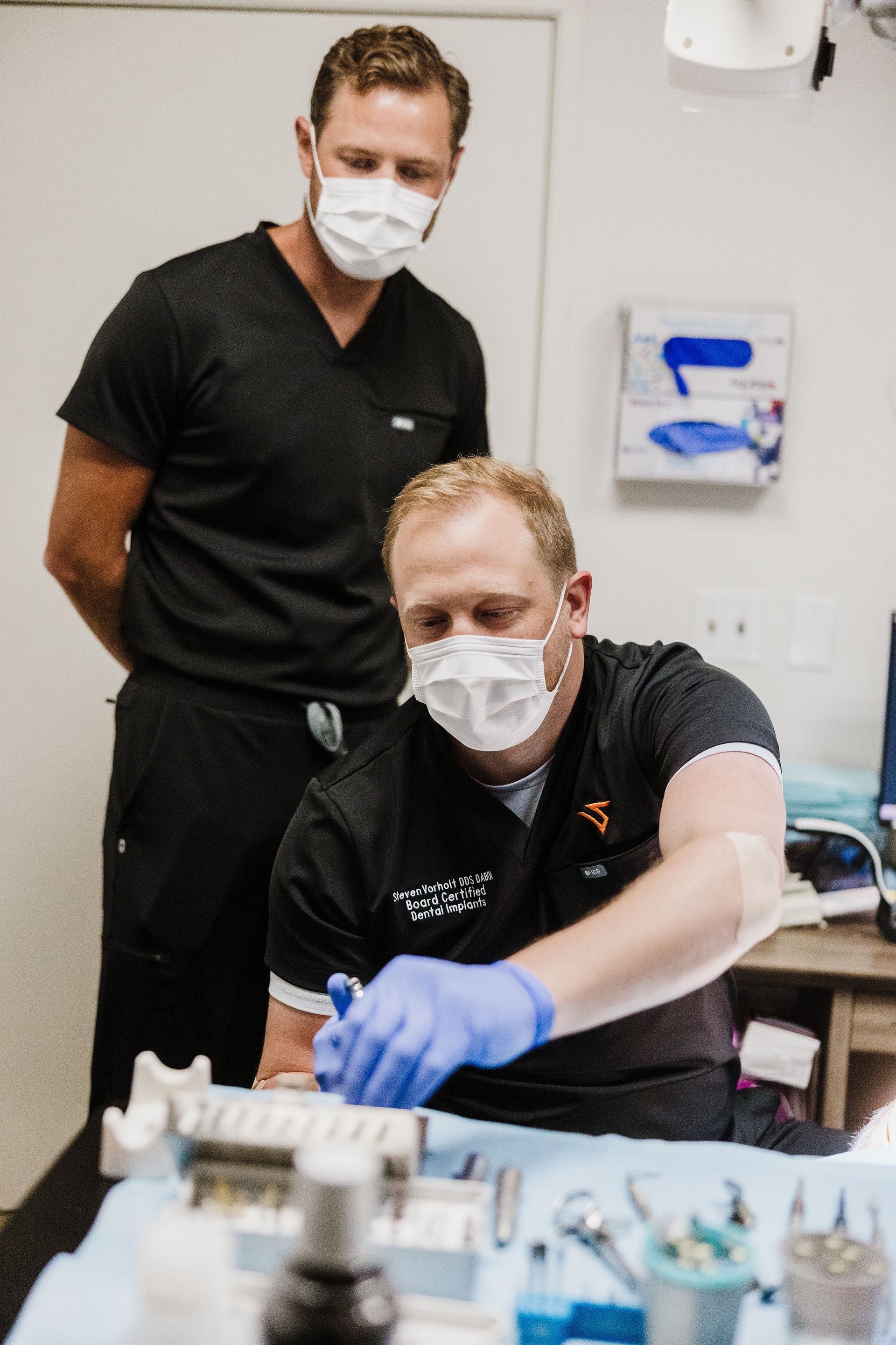 Dr. Aaron Miller looks over another dentist's shoulder in a clinic, both wearing black scrubs with a dental patient.
