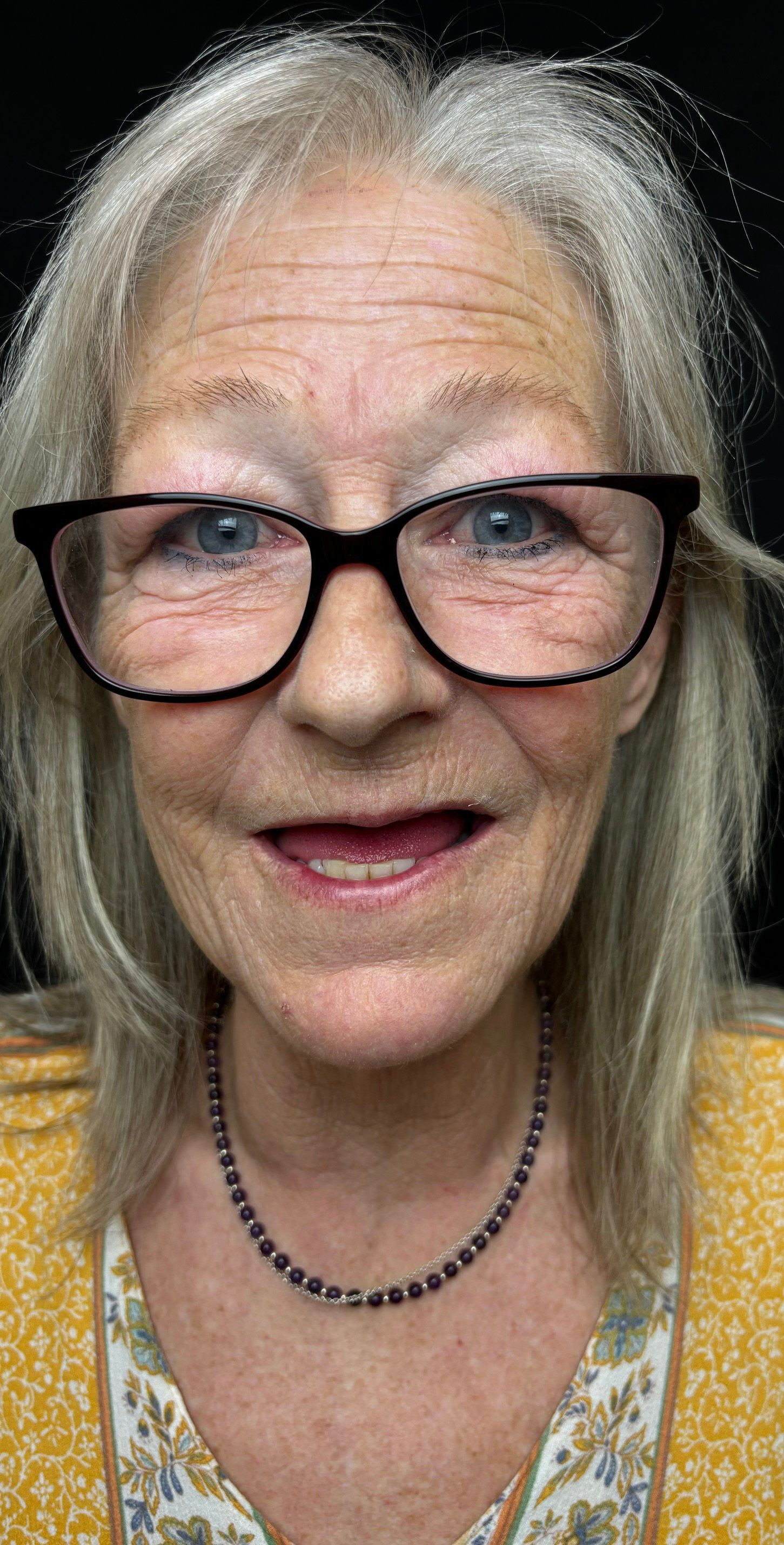 Close-up portrait of a smiling woman with glasses, gray hair, and a yellow top on a dark background