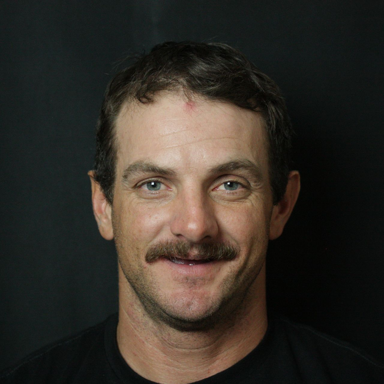 Smiling man with short dark hair and a mustache, wearing a black shirt against a dark background