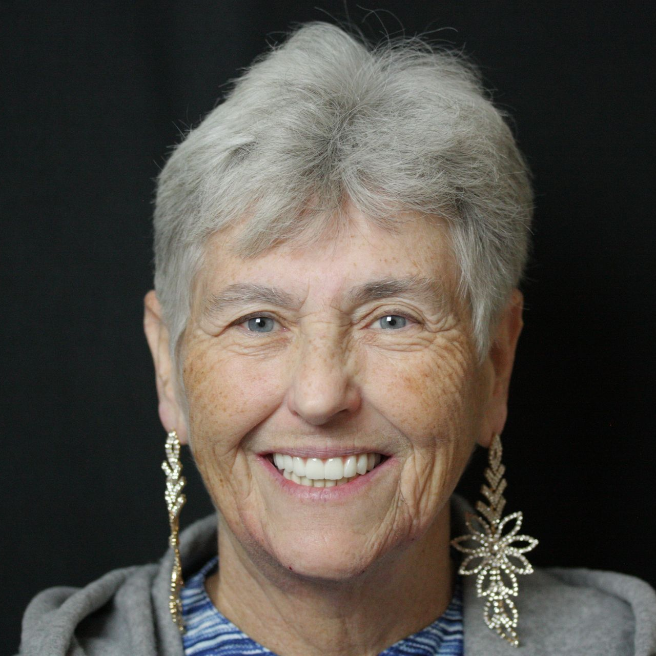 Smiling person with short gray hair wearing dangling earrings against a dark background