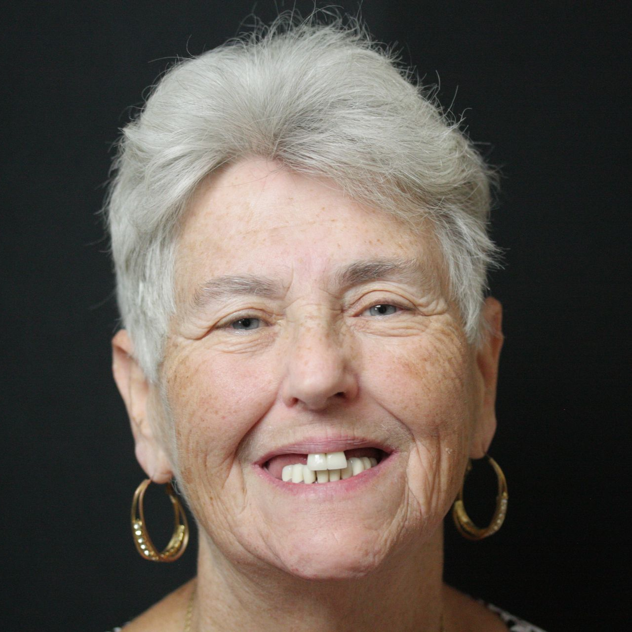 Smiling older woman with short gray hair and gold hoop earrings on a dark background