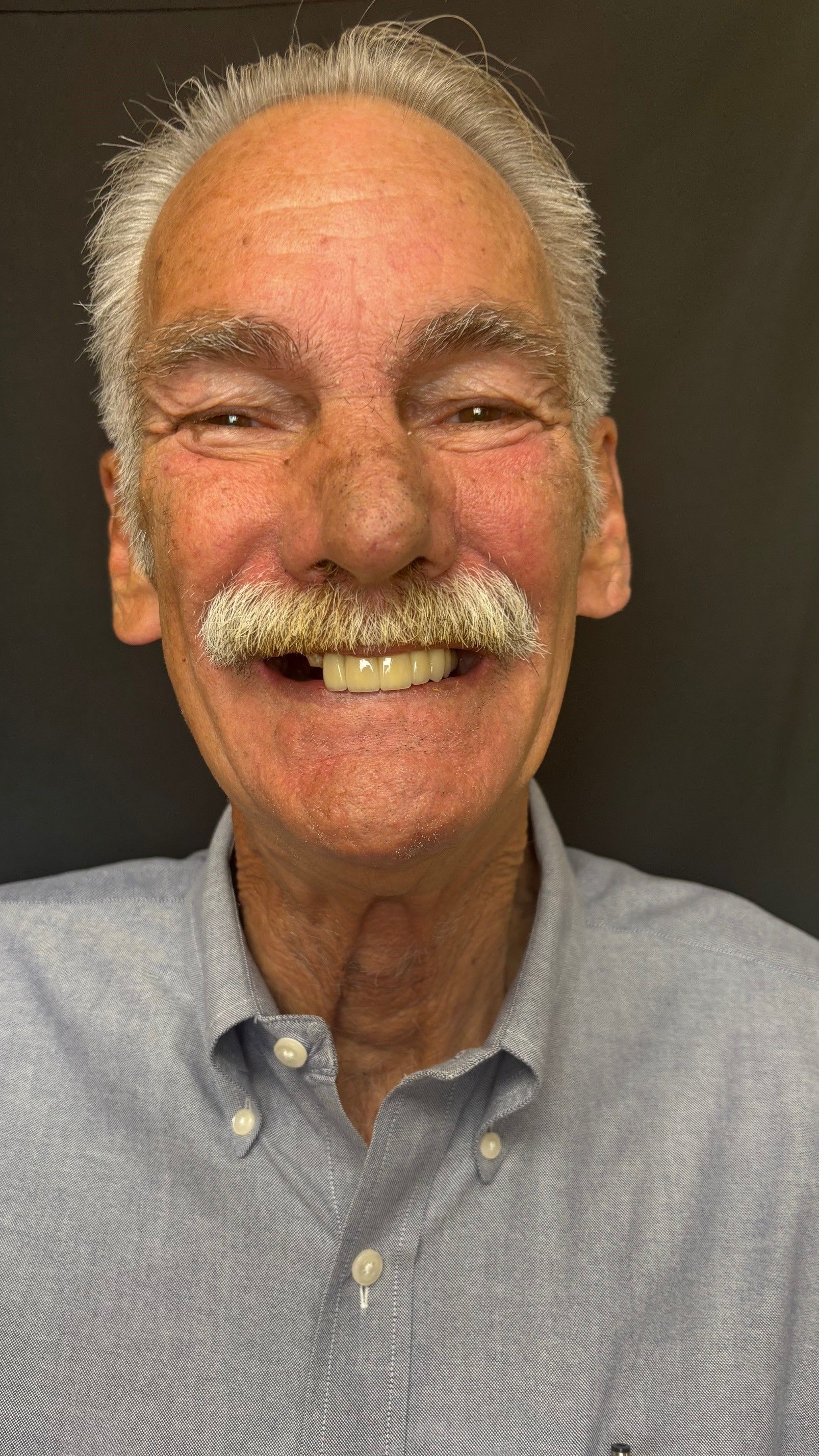 Smiling man with a white mustache, wearing a blue button-up shirt against a dark background