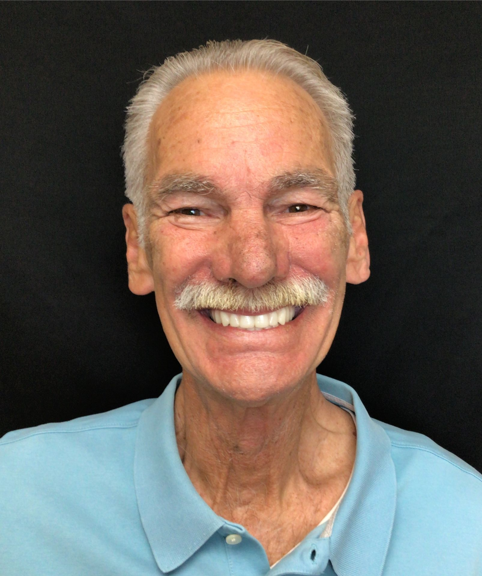 Smiling man with white hair and mustache wearing a light blue polo against a black background