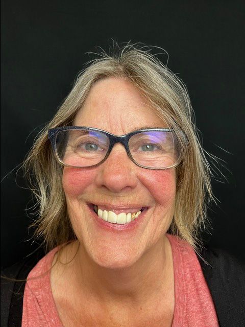 Smiling person with glasses and gray hair against a dark background