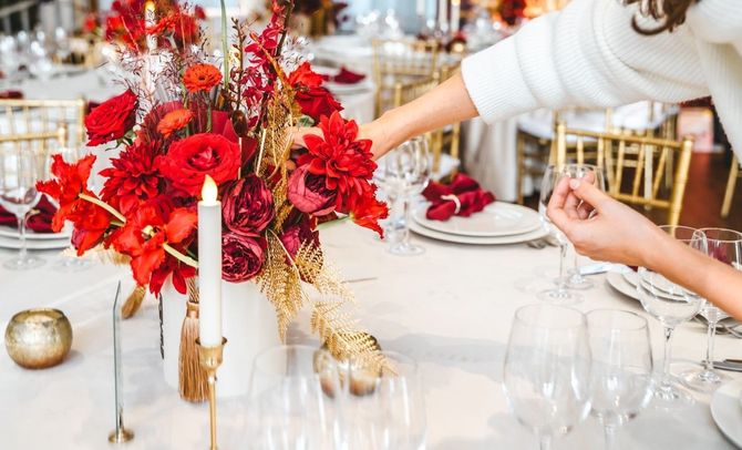 Elegant banquet table with red floral centerpiece, candles, and gold accents being arranged by a hand