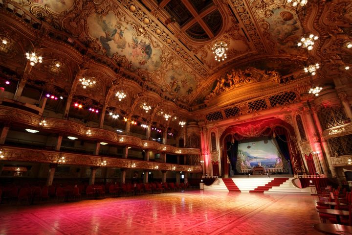 Ornate theater interior with red velvet seats, gold ceiling murals, and a lit stage
