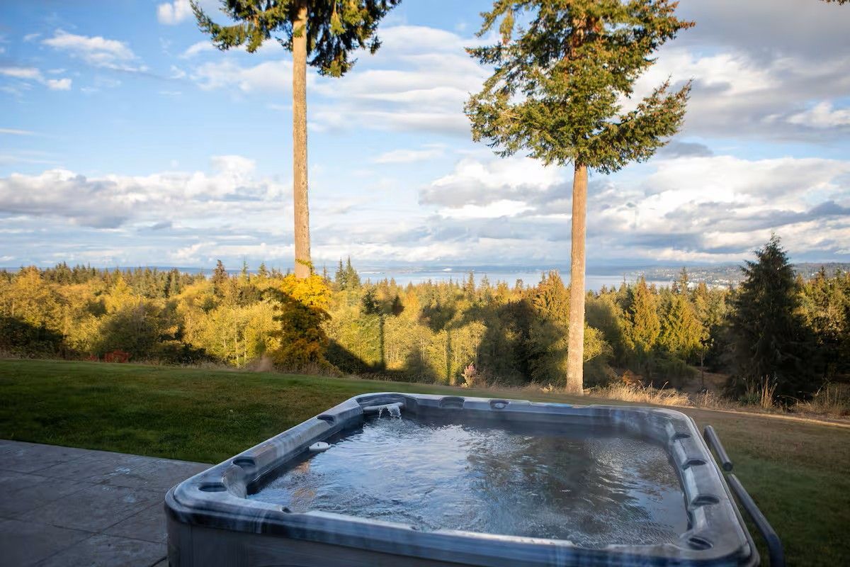 Hot tub on a patio overlooking a grassy yard, tall trees, and a distant mountain under a partly cloudy sky