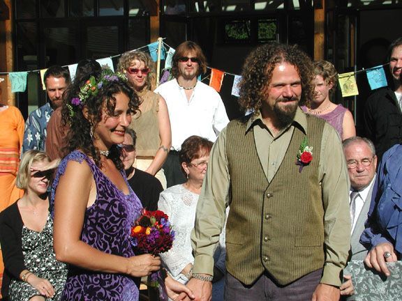 A couple holding hands at an outdoor wedding, with guests standing behind them.