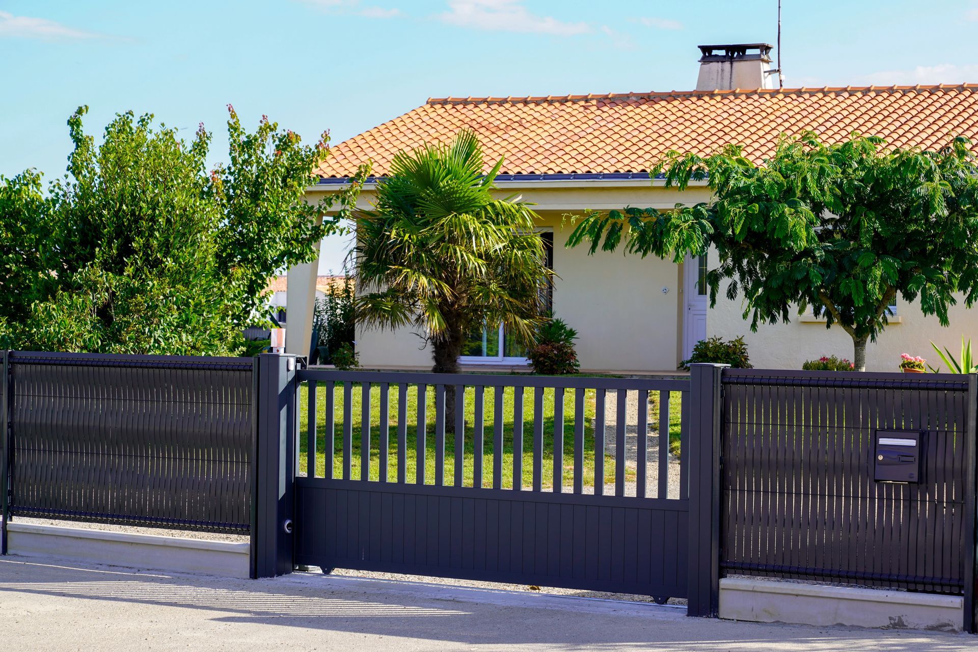 Casa con cancello e recinzione grigi, alberi e tetto di tegole arancioni in una giornata di sole.