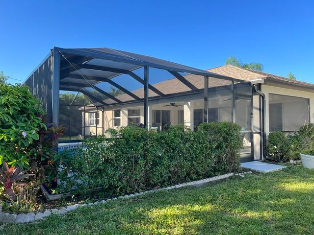 Backyard patio with black metal pergola, beige house, hedges, and green lawn in bright sunlight