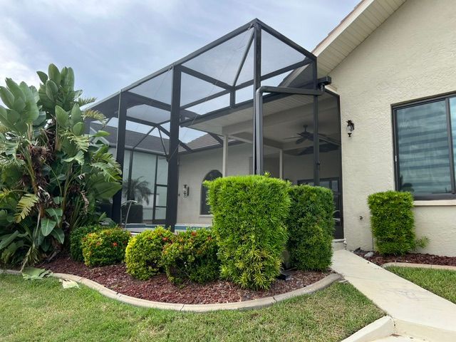 Patio with black screened enclosure beside a beige house, bordered by trimmed shrubs and lawn