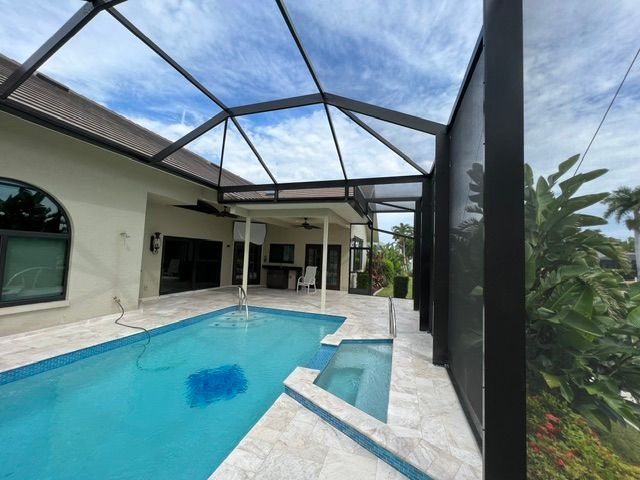 Covered backyard pool beside a patio and house, with black screen enclosure and lush greenery beyond