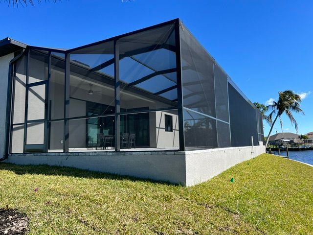 Modern glass-and-mesh pavilion on a grassy lawn beside palm trees and water under a blue sky