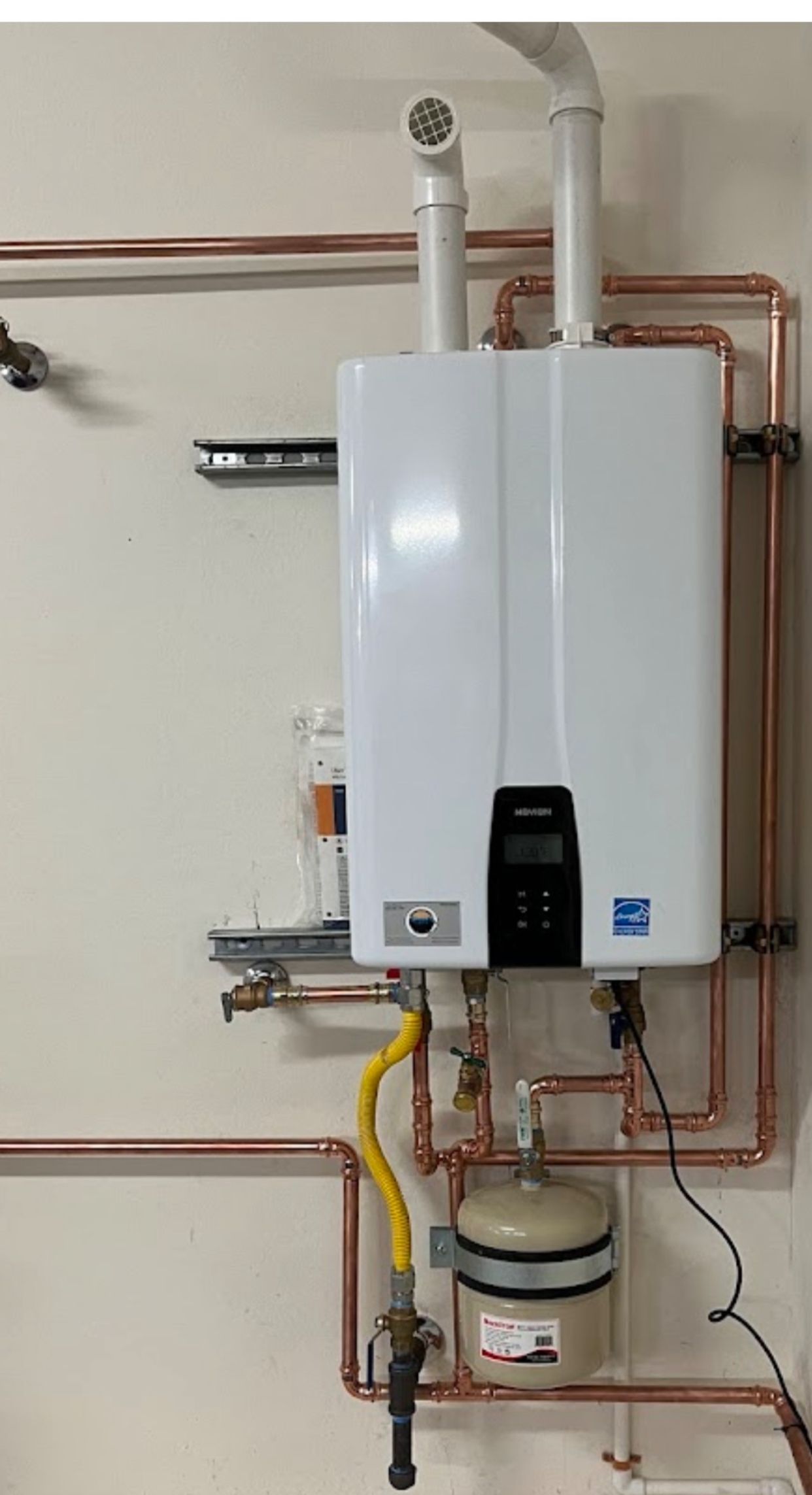 A white water heater is hanging on a wall next to copper pipes.