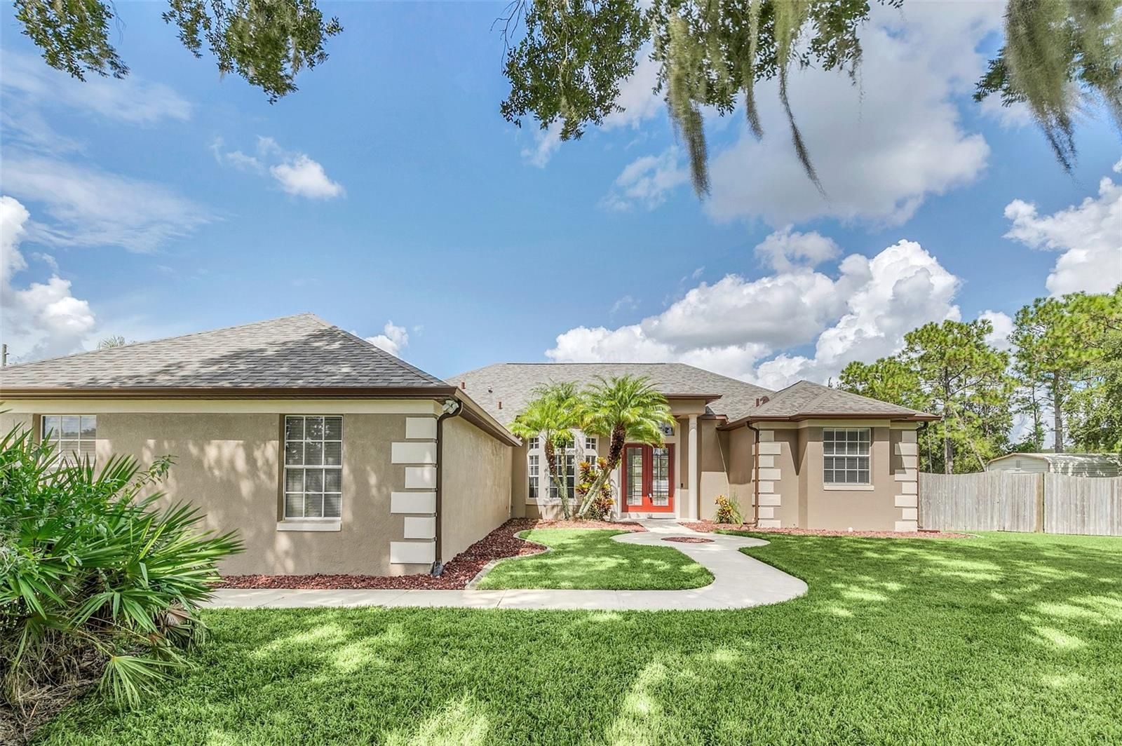 Tan house with white trim, red front door, and green lawn under a blue sky with clouds.