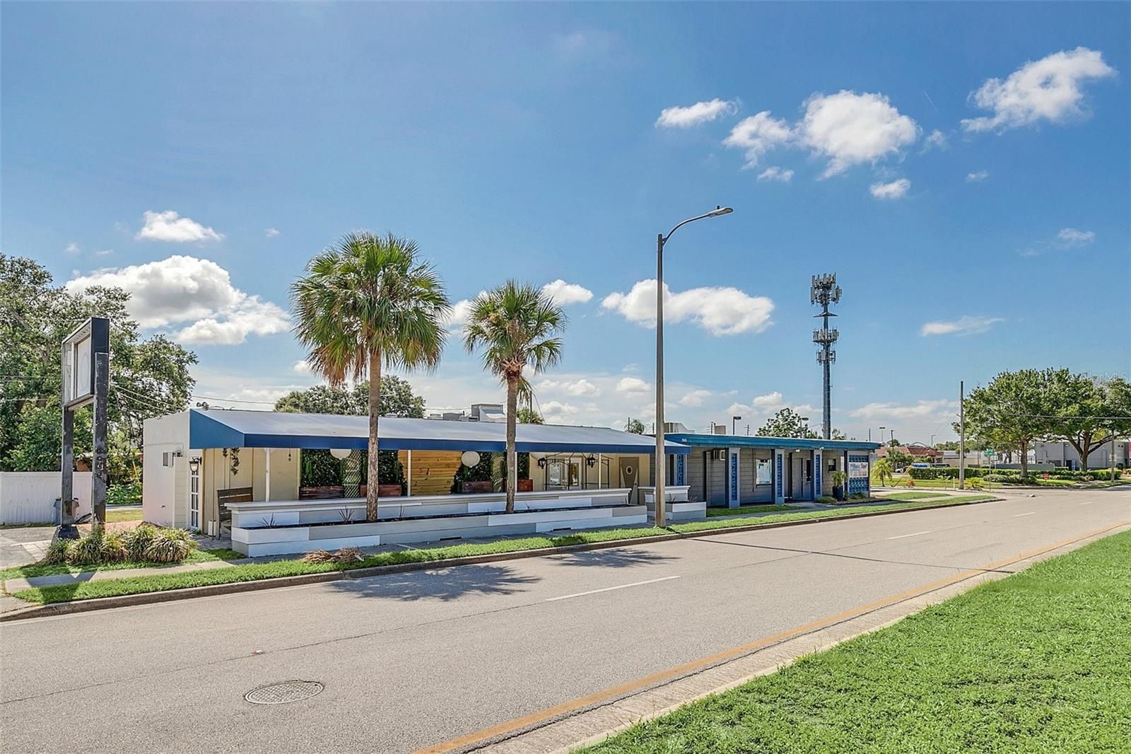 A long, low building with a blue roof and white walls, palm trees, and a street in front on a sunny day.