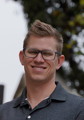 Man with glasses smiles outdoors, wearing a gray polo shirt, with a blurred background of trees and a building.