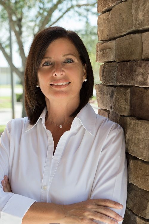 A woman in a white shirt is leaning against a brick wall and smiling.