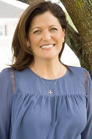 A woman in a blue shirt and necklace is smiling in front of a tree.