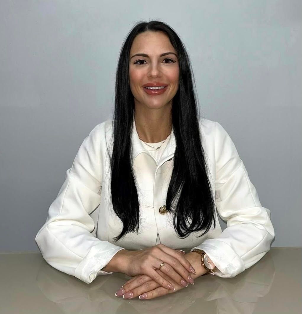 Una persona con cabello largo y oscuro, que viste una chaqueta blanca con cuello, está sentada en un escritorio con las manos entrelazadas.