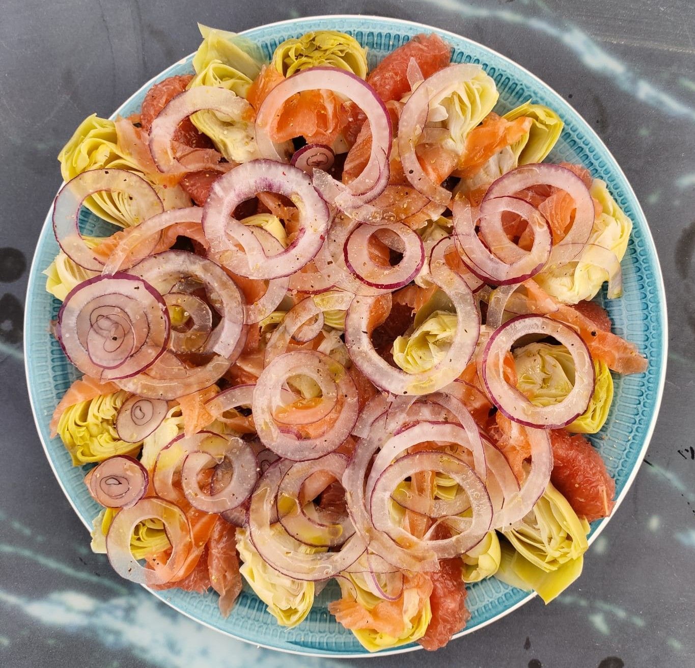Een salade van artisjokharten, gerookte zalm en rode uienringen op een bord met een blauwe rand.