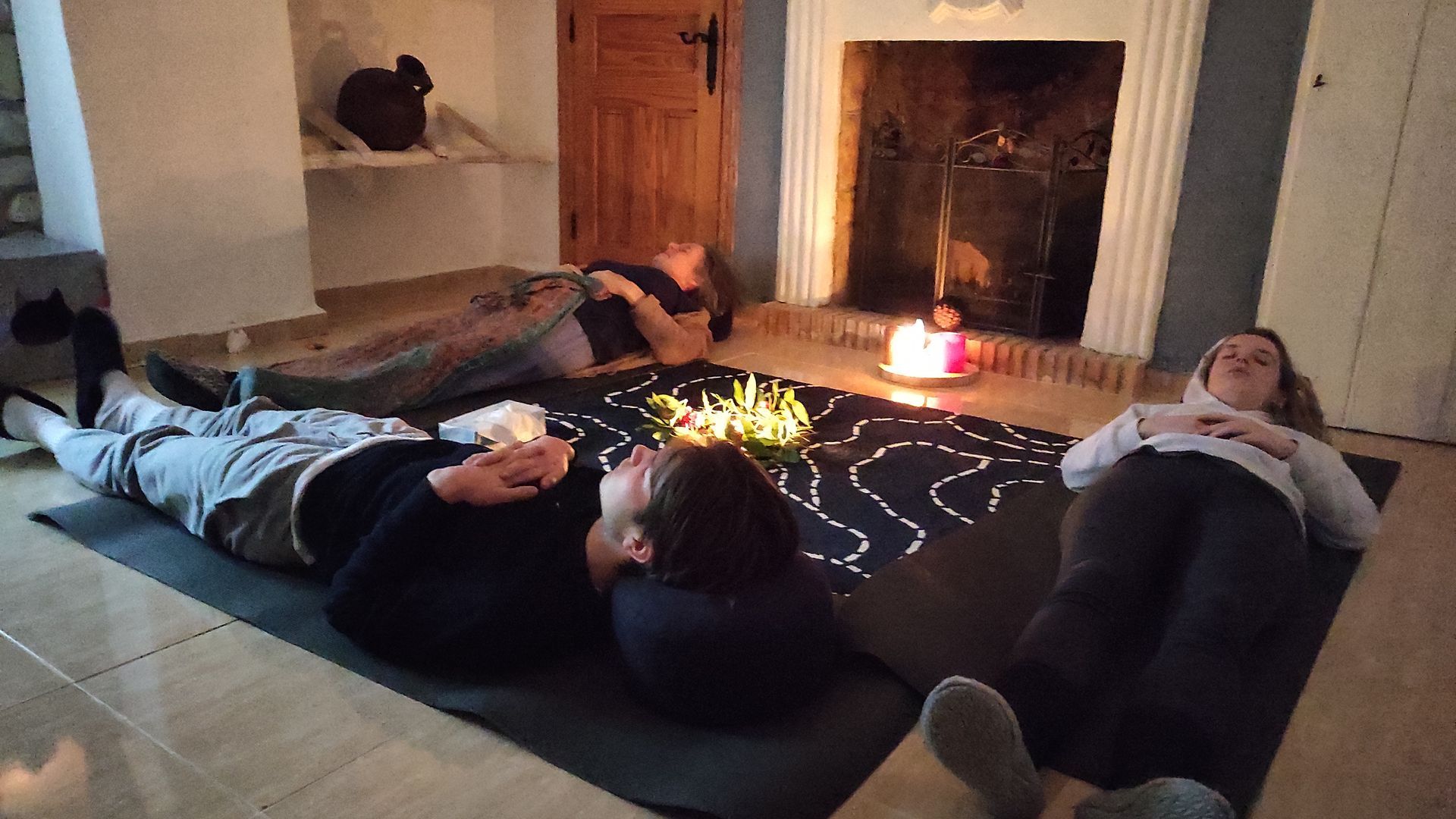 Drie mensen liggen op matten in een schemerig verlichte kamer met een open haard