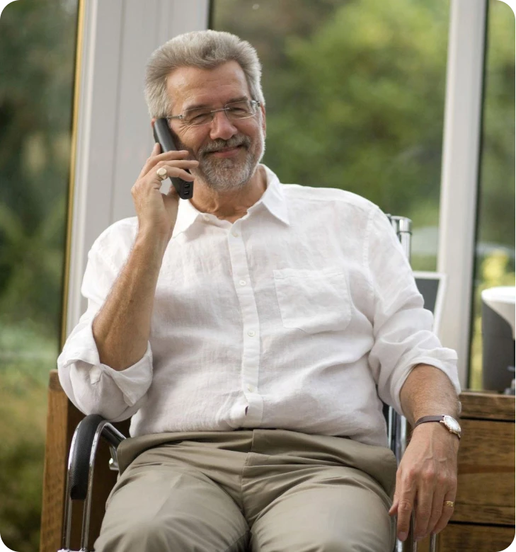 Man sitting on a wooden chair by a window, smiling while talking on a cellphone.