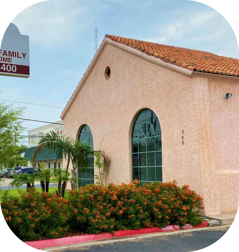 Small peach stucco building with arched green windows, red-tile roof, and flowering shrubs by the sidewalk