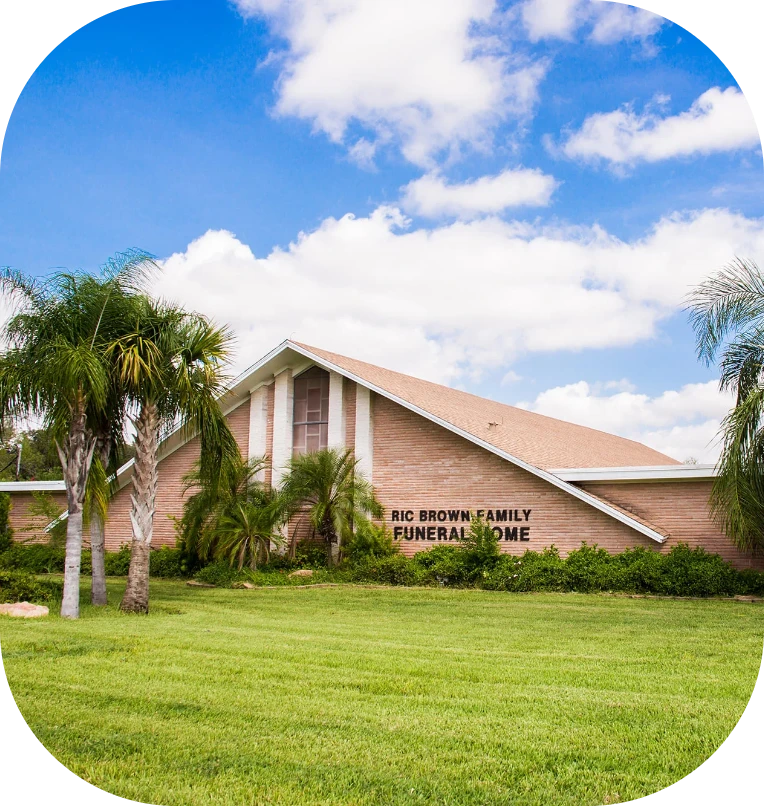 Brick funeral home building with a sign, surrounded by green lawn and palm trees under a blue sky.