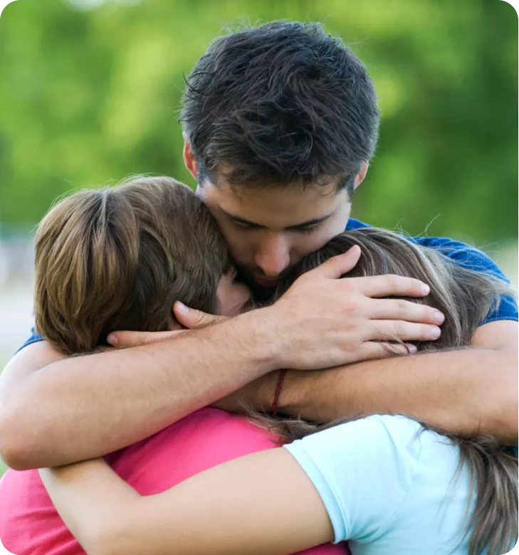 Three people in a close group hug outdoors, with green blurred background.