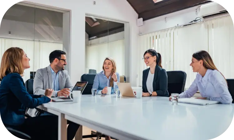 Five coworkers in a bright office meeting around a table, discussing with laptops open