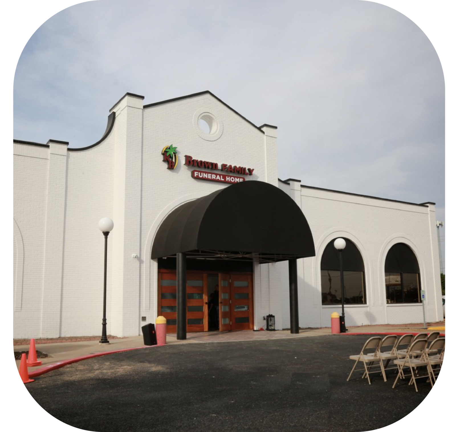 White building with black awning and arched windows, seen from the parking lot with folded chairs outside.