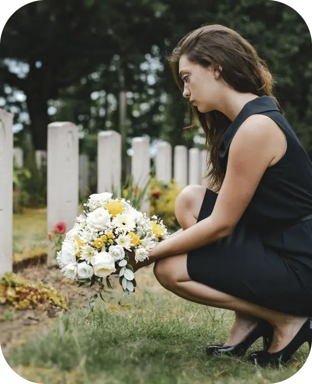 Person kneeling at a gravesite, placing a white and yellow flower bouquet by headstones.
