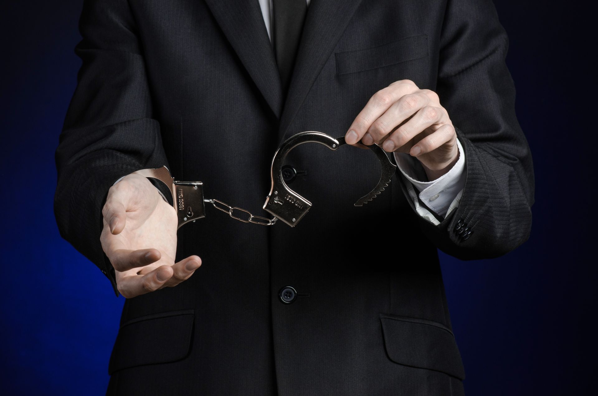Person in a black suit holding open handcuffs against a dark blue background
