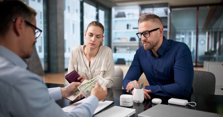 Three people in a modern office discuss documents across a table with coffee mugs.