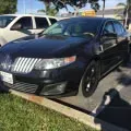 Black sedan parked on a grassy curb beside a sidewalk, with another car and palm trees in the background
