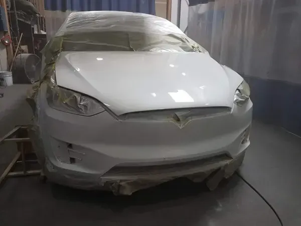 White car with front-end damage in a body shop, hood crumpled and covered with masking film.