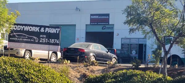 Black car parked outside a bodywork and paint shop with a white building and trees nearby