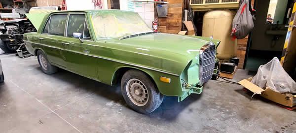 Green classic car in a cluttered garage, front end partially disassembled for restoration.