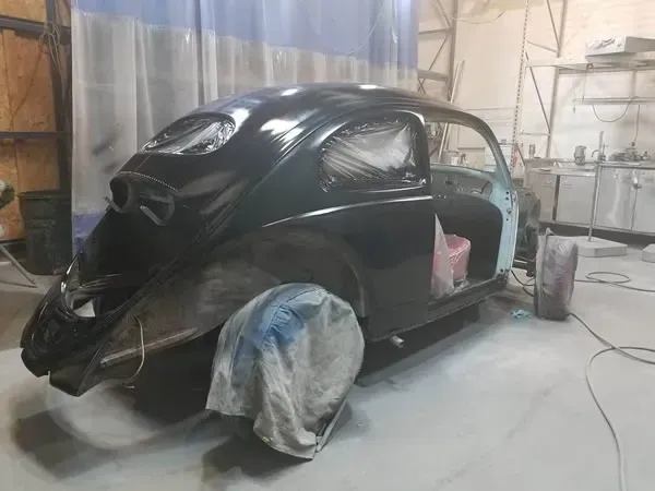 Black classic car body shell in a workshop, partly covered with tarps and missing wheels.