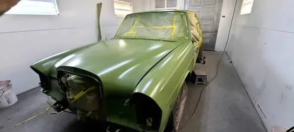Primer-green car body in a narrow garage, front end missing and windows taped off for restoration.