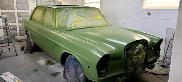 Green classic car in a garage, covered in masking tape and ready for painting.